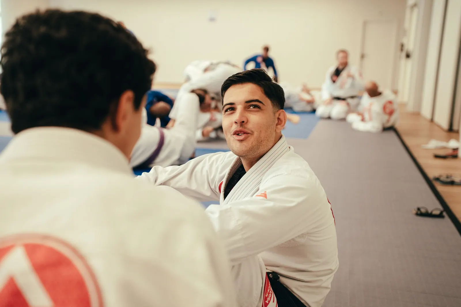 A student in a white gi talking with a teammate on the mats